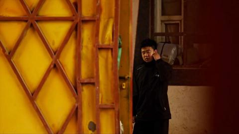 A man in a black jacket holds his ear and watches on as the camera films