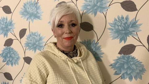 A woman with short cropped bleached hair has bright red lipstick and is wearing a cream hooded top. She is standing in front of wallpaper with large blue flowers on a cream background