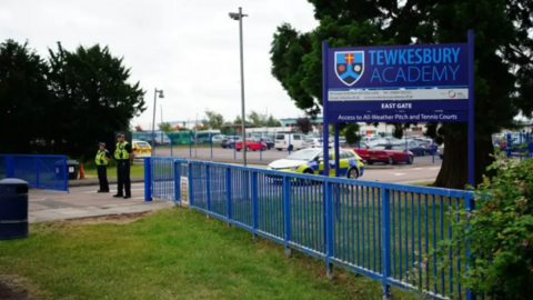 The front gates and fence at Tewkesbury Academy, which are blue, as is school sign and crest. There are two police officers stood at the open gate and a police car preparing to drive out of the school grounds.