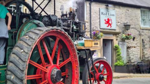 A green traction engine with red wheels outside a rural pub with a sign saying "The Red Lion"