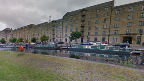 A canal with several small barges and boats moored there. There are flats and cars alongside it