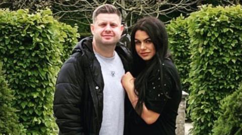 Josh and Chloe Bashford standing together in front of a hedge looking to camera