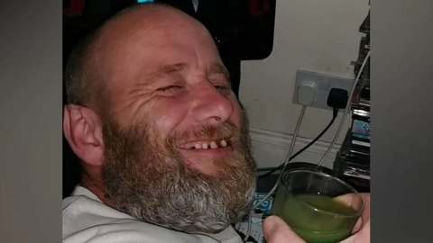 A close-up photo of David Hunt's face. He is smiling at the camera and wearing a white T-shirt while holding a glass of green liquid