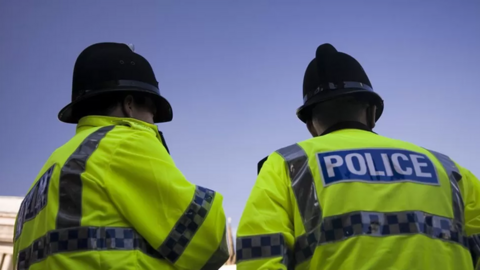 Two police officers in hi-vis jackets and black police hats stand together in front of a blue sky