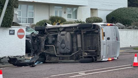 An overturned van in Marine Gate, Brighton. The underside of the silver van can be seen in the image. Part of the bumper has fallen off the van. 