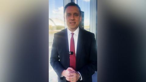 Anas Sarwar smiles at the camera as he stands in front of a wide window. He has black hair and brown eyes and is wearing a red tie, white shirt and black suit.