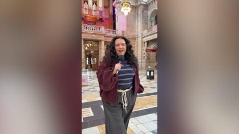 Ashleigh Storrie stands in a museum holding a microphone. She has long, dark hair.