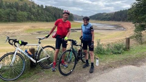 A man in a red cycling jersey and shorts and a woman in a blue cycling jersey and shorts stand beside their bikes in front of an empty reservoir.