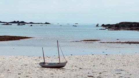 A small boat on the sand with pale blue sea behind and a pale blue sky behind 