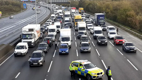 A police car blocks the flow of traffic across six lanes of the M25 