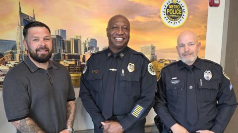 Taylor Johanson stood with John Drake and Peter Kinsey in front of an image of the Nashville skyline. Drake and Kinsey are in navy police uniforms while Johanson is in casual clothes. All three men are smiling.