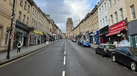 A photograph of Park Street, with the Wills Building at the top of the road in the centre. The sky is cloudy and the road is empty apart from a few parked cars. There are shops lining the street including a homewares store and a vintage clothes store.