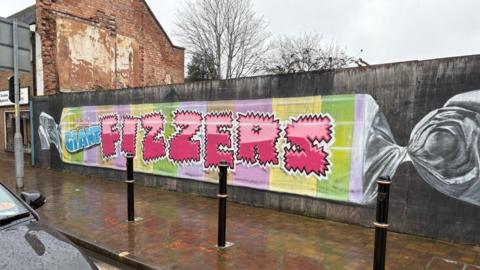 A long wall beside a wet sidewalk shows a mural painted like a wrapped candy. It has pastel stripes and large pink letters spelling “FIZZERS.” 