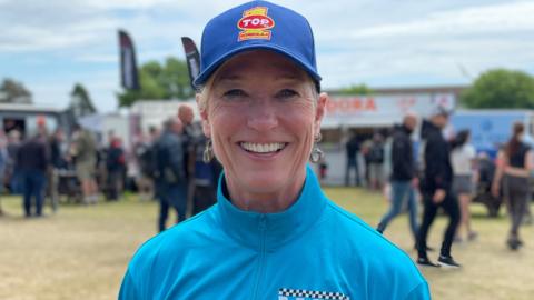 Erin Sills has short blonde hair and is wearing a dark blue cap and lighter blue sports top with a zip up to the neck. She is standing outside with groups of people walking around behind her.