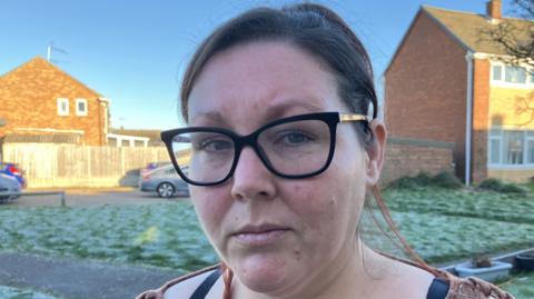 A woman with black hair and wearing black rimmed glasses and a brown top standing in front of a grassy area with terraced houses in the background. 