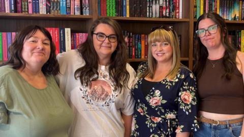 Carrie McGovern, Sara Mack, Ellie White and Hayley Beckinsale. Carrie is wearing a sage green dress. She has shoulder, length brown hair that is curled, with side bangs. Sara has long brown hair that is curled. She is wearing black-rimmed glasses, and a white top that has a design with three mountain peaks within a circle, and the word Velaris above them. Ellie has blonde hair that is medium length with a fringe. She is wearing a floral dress that is navy with pink peonies and roses, and she has a pair of tortoiseshell sunglasses on the top of her head. Hayley has long, dark brown hair, and is wearing tortoiseshell glasses and a brown vest top. They are stood together in front of a bookcase. Each shelf is full with an array of books in different colours and genres. 
