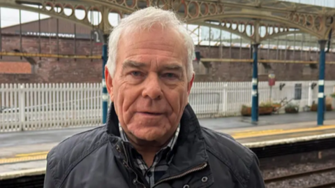 Ken Harper stands at a train station. He has short white hair.