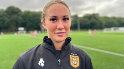A girl wearing a Hull City Ladies black quarter zip with an Orange Hull City badge on, she has blonde hair and long eyelashes and she's smiling at the camera