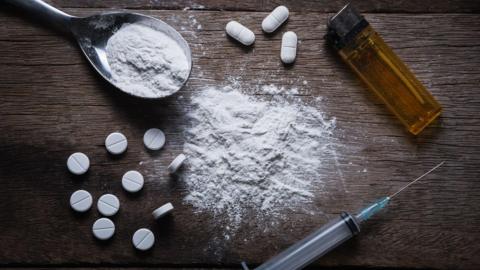 A brown table, on which several white pills are scattered alongside a lighter, a syringe and a spoon full of white powder. There is more white powder on the table.