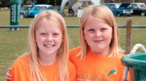 Jemima and Isabel standing arm in arm wearing orange T-shirts. They are standing behind a table at a fundraising event and there is grass in the background.