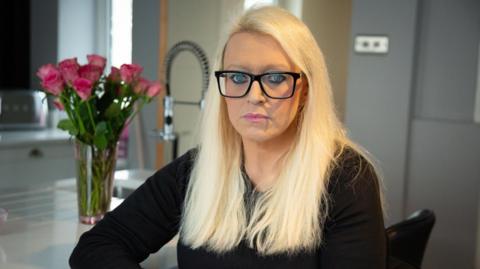 Betsan, who has long bleach blonde hair and glasses and is wearing a black long-sleeved top. She is sat at a kitchen counter, with a sink and a vase of pink flowers beside her and grey cabinets in the background.