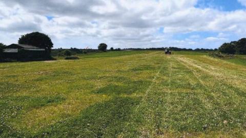 The view down a grass airstrip. To the left is a barn and some trees. In the distance is a tractor mowing the grass.