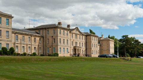 The outside of the hospital, a large Georgian building with columns either side of its front door. It has large bay windows and green lawn to its front. There are a few cars parked outside its front door. 