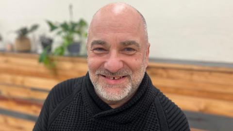 Simon Berry is wearing a black jumper and he is smiling at the camera. He has short white hair and a closely shaved beard. He is sat in front of a wooden shelf with plants on it.
