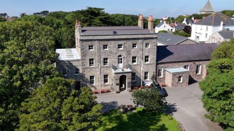 A drone shot of Island Hall. It is a Georgian-style building with grey bricks, and a large porch entrance.