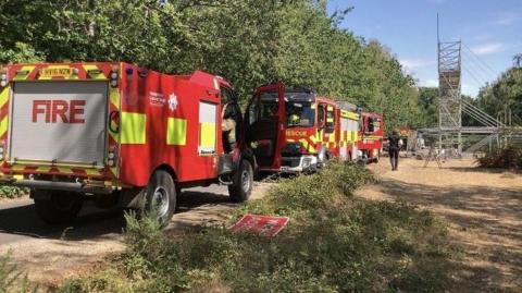 Red fire and rescue vehicles line a tree-bordered lane leading to a scaffold tower and fenced work area in a woodland setting. There is a person in dark uniform in the distance.