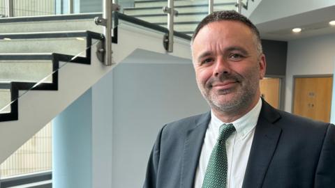 A man in a suit with a white shirt and green tie poses within a college building