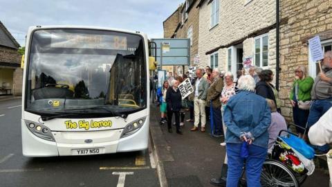 People campaigning with placards as the 84 bus arrives at a bus stop. The bus is cream coloured with yellow writing on the front