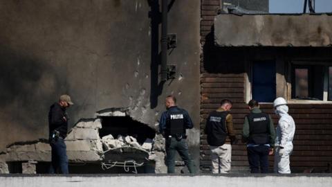 Four men in police vests, as well as a man in a white hazmat suit, a helmet and a face mask, are seen stood next to a building which has a hole in its side following the drone hit