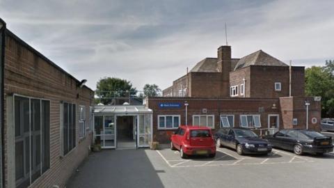 The outside of a brick hospital, with cars parked and a blue Main Entrance sign.
