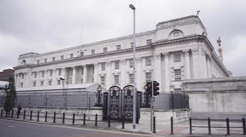 The High Court in Belfast. It is a white building with large back gates. It is situated on a main road beside a pedestrian crossing.