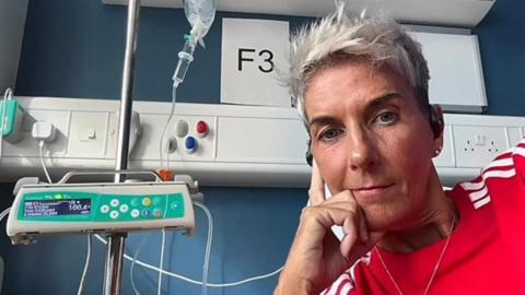 Carmel is looking at the camera and is leaning against her hand. She is in a hospital setting and wires and tubes can be seen beside her. She is wearing a red top with white stripes and has short grey hair. 