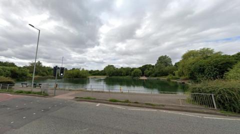 A body of water, behind a pedestrian crossing, is surrounded by trees. There is railing in the foreground and a streetlight.