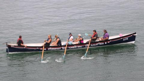 A long gig boat in a harbour with seven rowers on it. The boat is navy and has a white interior, as well as a white stripe and a red stripe towards the top of it. It has some words on the side which read 'StormFest 160 - Torbay RNLI - 22nd August 2026'. Six of the rowers are holding oars, which are light brown with teal coloured ends.