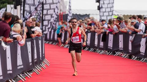 A man wearing a vest and shorts sprints down a red carpet for a sporting event. Crowds cheer him on from the sidelines.
