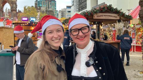 Two women stood smiling next to each other. There are wooden market stalls behind them, and they both wear a red and white Santa hat. 