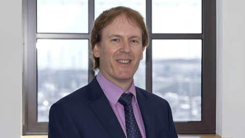 A picture of a man dressed in a suit looking straight at the camera. He is wearing a purple shirt with a blue blazer and blue spotty tie. He is stood in front of a window and is smiling. 