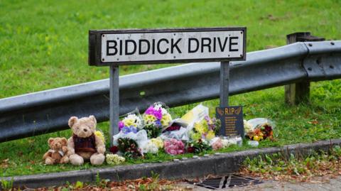 Close-up of a street sign, Biddick Drive, near the scene of the Keyham shootings. The horizontal sign on two metal legs is in front of an Armco safety barrier with grass in the background. At the foot of the sign, running along the kerb, are two teddy bears, numerous bouquets of flowers in plastic wrappings, and a small plaque headed "RIP".