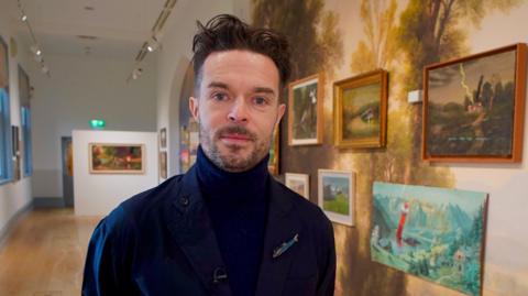 Oliver Jeffers is a man with black hair and beard who is standing in an art gallery in front of a wall of artworks. He is wearing a navy turtleneck top and coat. 