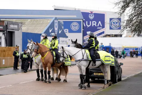 Four police officers on horses are in front of Fratton Park stadium in Portsmouth, other officers stand behind them closer to the ground