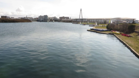 A wide shot of the lake. There is grass on the banks and canoes resting there.