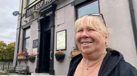 A woman with blond hair and black glasses perched on top of her head standing outside a pub. 