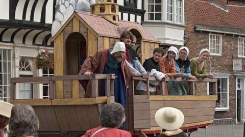 Seven people stood on a raised platform re-enacting a play. They are dressed in Medieval clothing.
