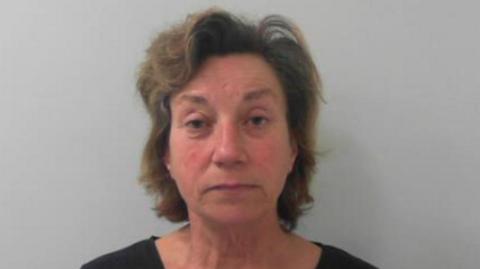 A police mugshot of a woman aged around 60. She has fairish shoulder-length hair, a red face and is wearing a black top.