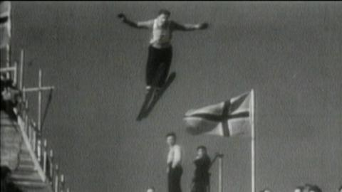 A black and white photo of a ski jumper taking off and flying through the air, with his arms outstretched. A Norwegian flag is fluttering on a flagpole behind him.