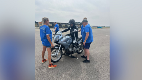 A picture of Ted Spurrell sitting on his specially modified motorbike. Ted is wearing a black helmet and black outfit. Ted is being helped by three members of staff wearing blue t-shirts.